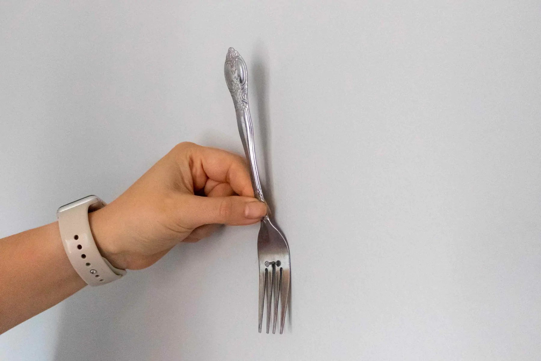 A hand holding a stainless steel fork against a white wall with the tines curved toward the wall, positioned to guide a picture wire onto a hanging nail.