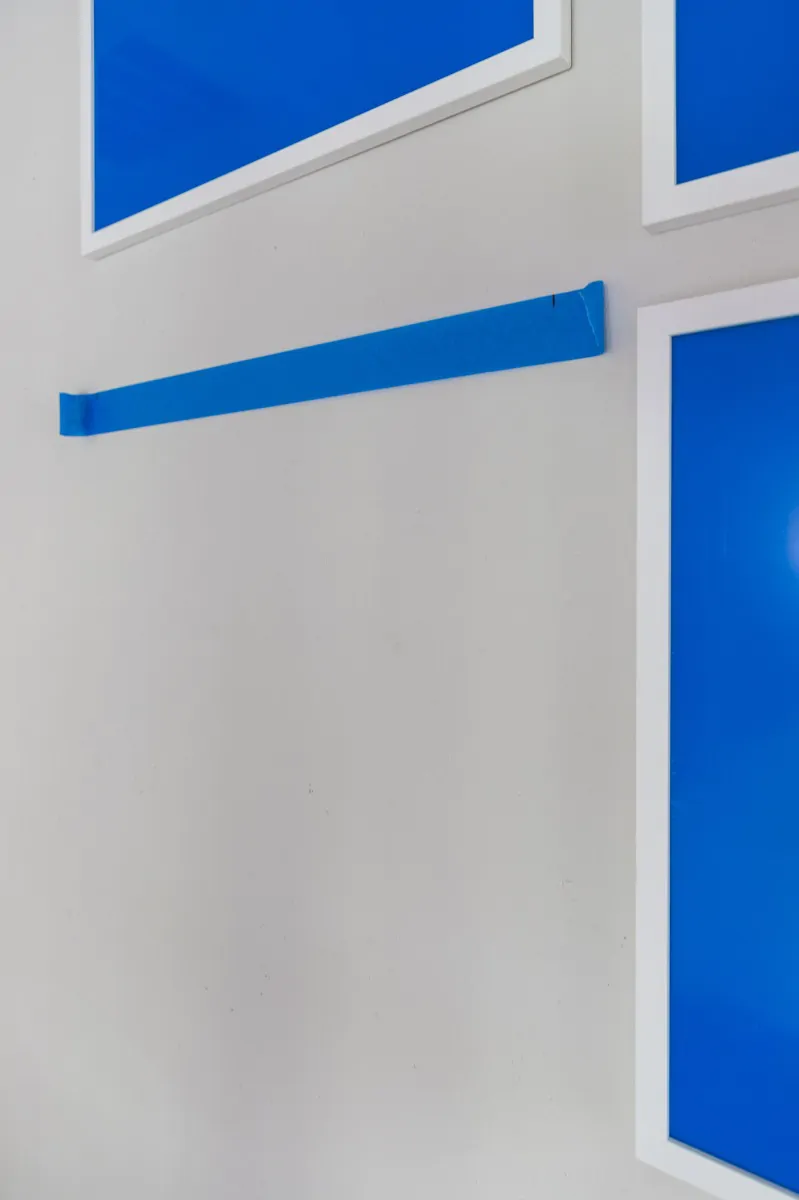 A horizontal strip of blue painters tape stretched across a white wall between three hung blue prints, with the transferred hanger marks visible on the tape.