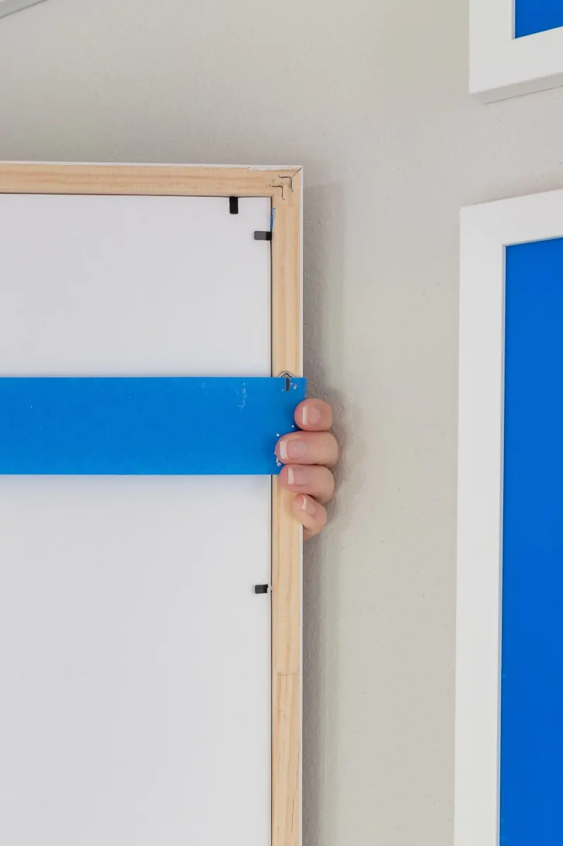 A strip of blue painters tape pressed across the back of a wood picture frame, capturing the position of the picture wire and both hangers along the top rail.