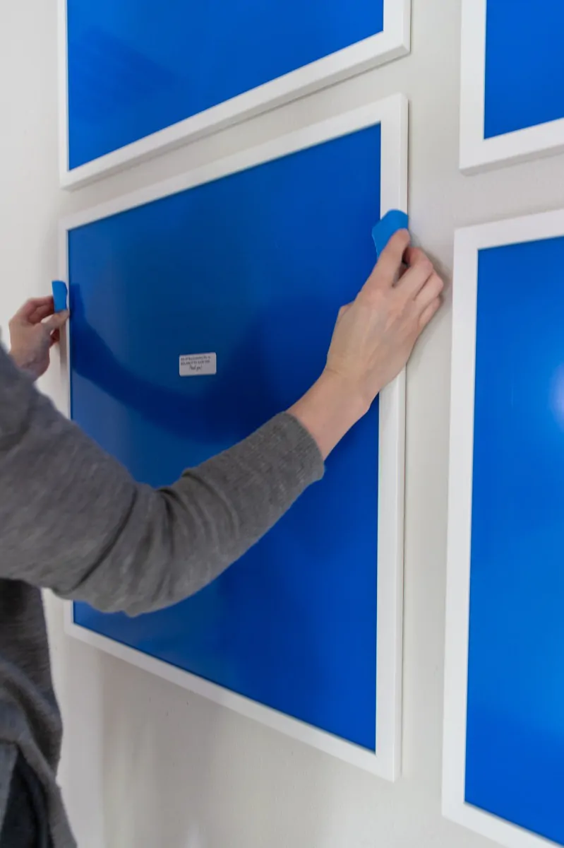 A hand placing a small blue painters tape tab at the corner between framed blue prints in a four-frame gallery grid, marking the corner position for the next frame before drilling.