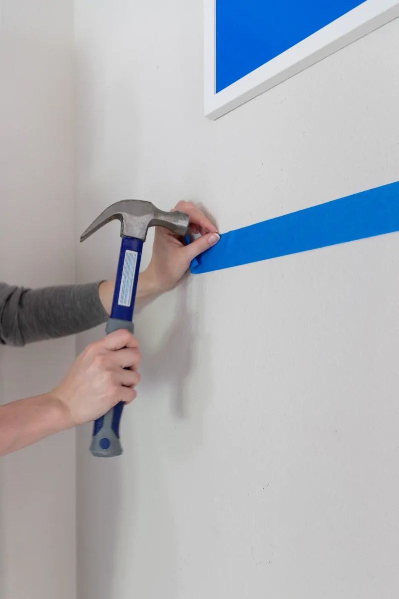 A small hammer driving a hanging nail through the marked painters tape on the wall, with a finished frame already in position above.