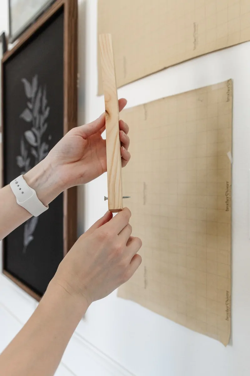 A hand holding a length of pine scrap with a small screw poking through one end, ready to be used as a transfer tool for marking a sawtooth hanger position on the wall.