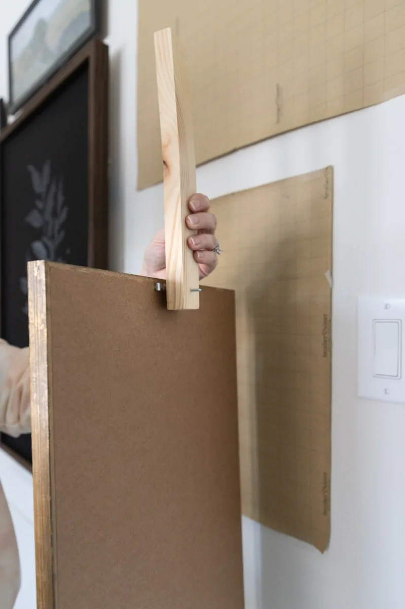 The wooden marker stick aligned along the back of a picture frame, with the screw tip lined up against the sawtooth hanger to record the offset between the hanger and the top of the frame.