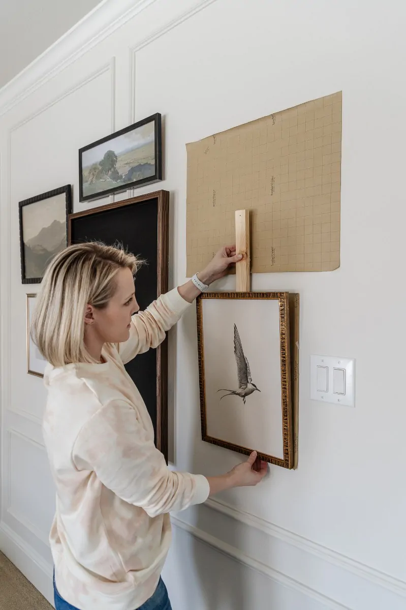 A person pressing the marker stick against a kraft paper template on a paneled wall to set the nail spot for a framed bird sketch in a gallery arrangement.
